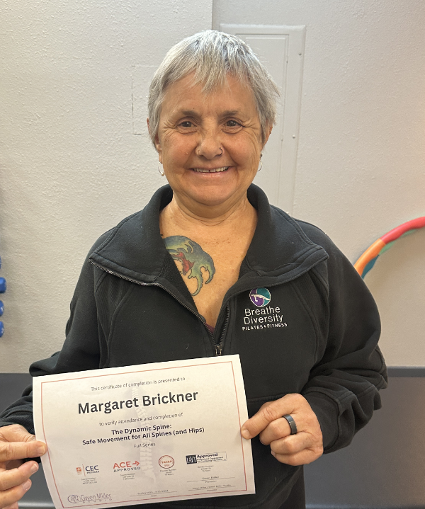 Smiling woman holding a certificate, wearing a Breathe Diversity jacket and showing a colorful chest tattoo